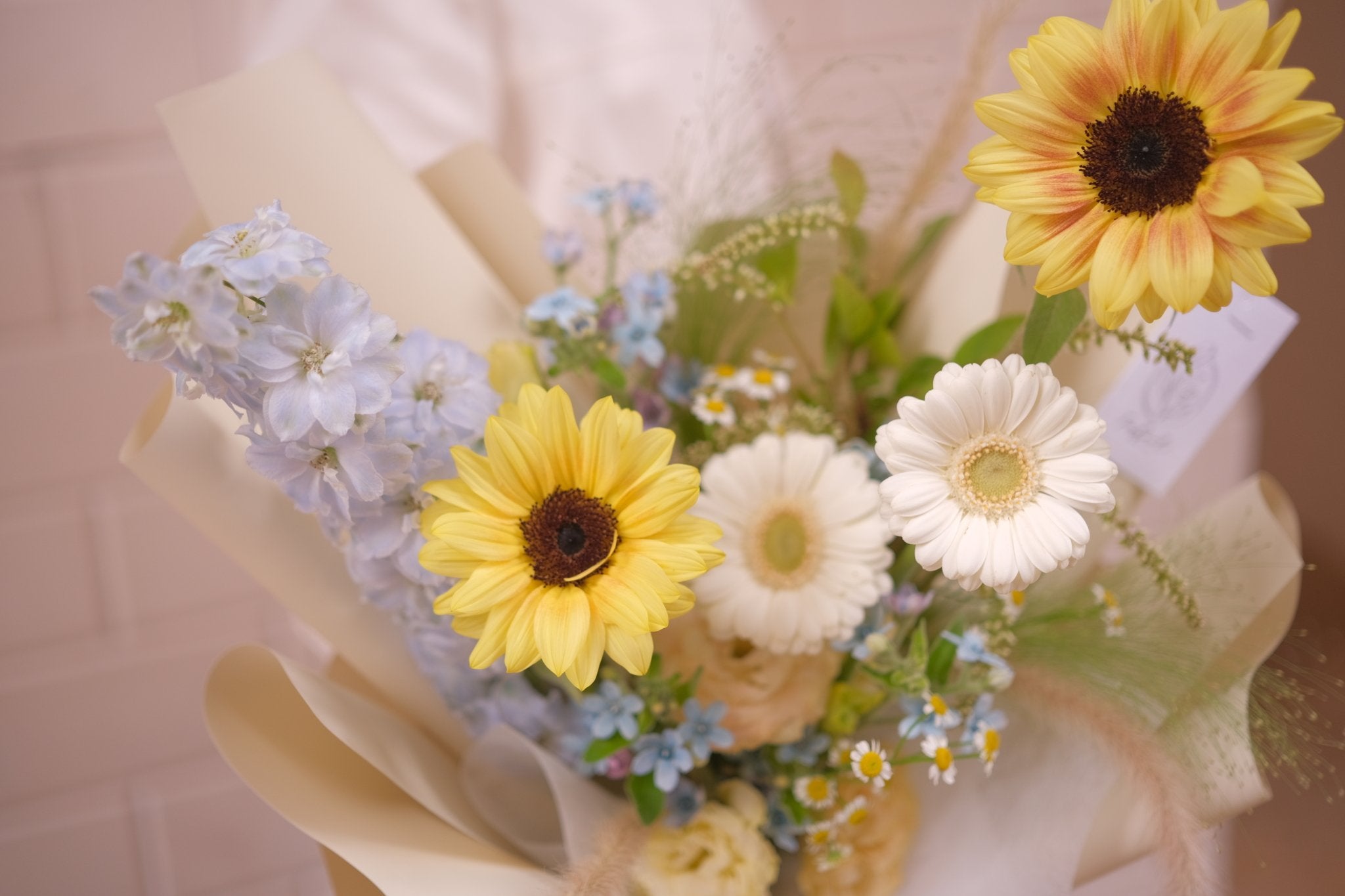 Yellow sunflower graduation bouquet with white daisies and blue accents in beige wrapping, perfect congratulatory surprise for graduates.