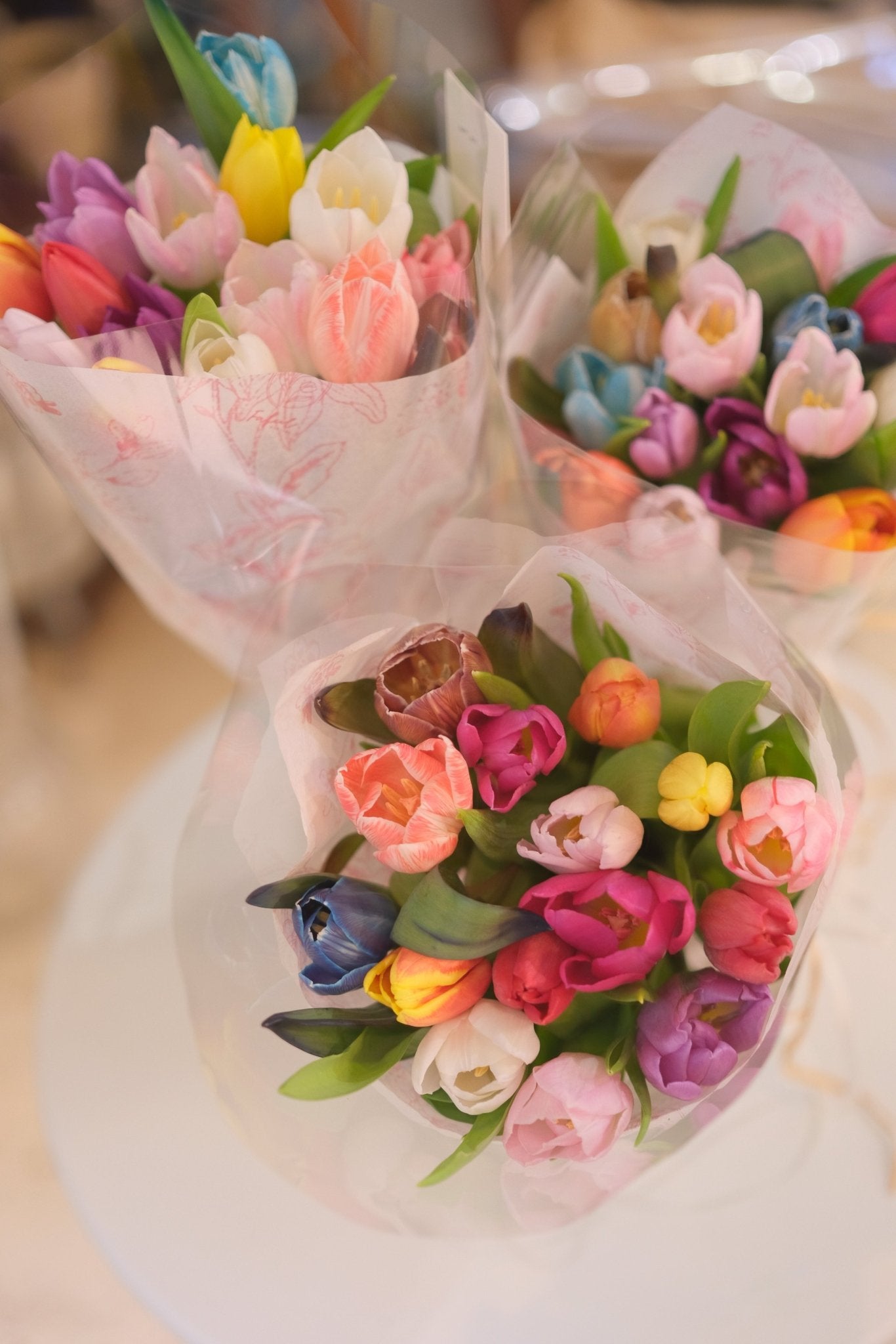Three pastel-wrapped rainbow tulip bouquets with dense rounded blooms on light surface by Faful Florist
